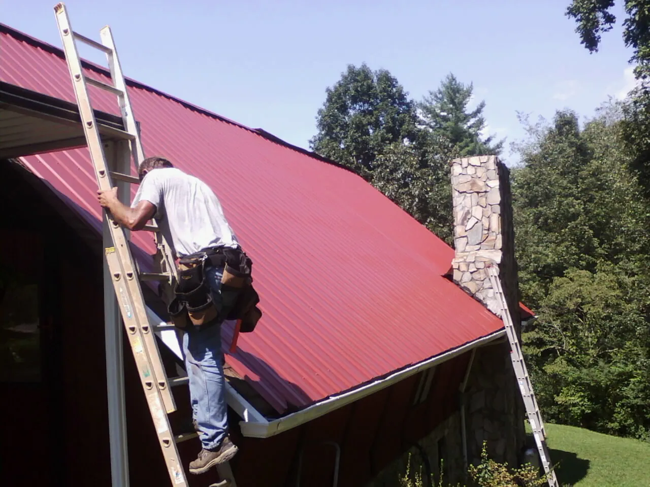 Roofer on a ladder beside a red metal roof and stone chimney — Soffit & Fascia Repair by Roofing Brothers in Crofton